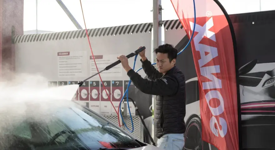 Man using a pressure washer on a car windshield, with a SONAX banner in the background.