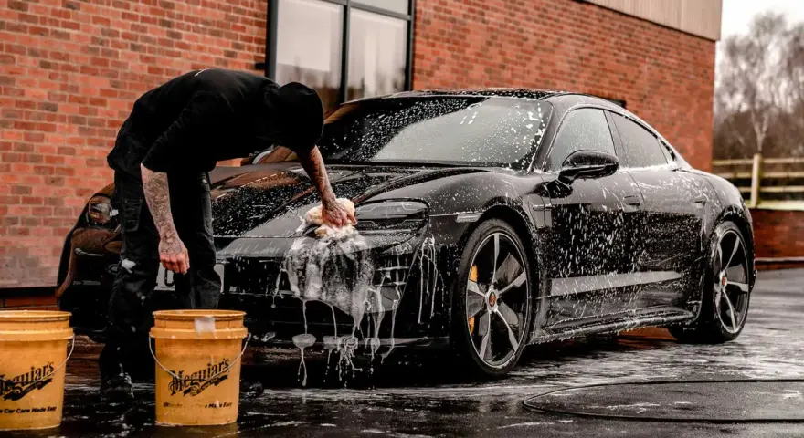 Person tvättar en svart bil med Autoglym-produkter. Två gula hinkar står bredvid.