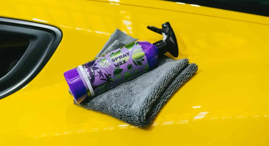 En flaska spray wax ligger på en gul bil med en grå mikrofiberduk.