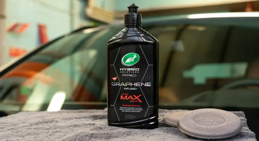En flaska Turtle Wax Hybrid Solutions Pro Graphene Infused To The Max Wax står på en grå trasa bredvid två appliceringsdynor.