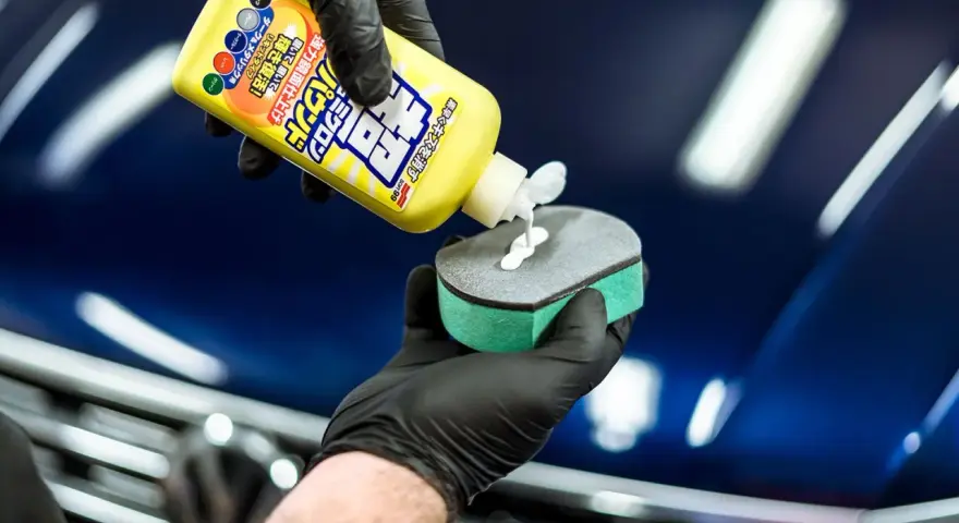 En hand med svart handske applicerar en vit kräm från en gul flaska, märkt Soft 99, på en svamp för bilvård.