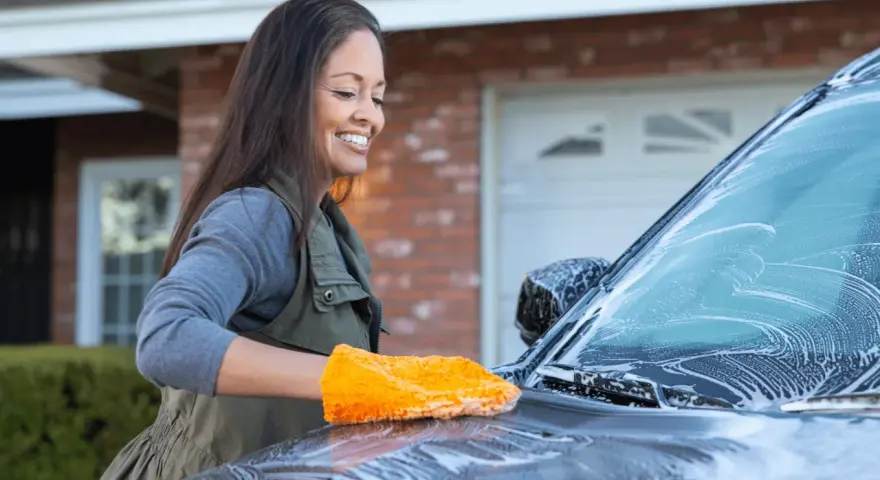 Glad kvinna tvättar bilen med en orange svamp. Hon putsar bilrutan som är täckt av skum.