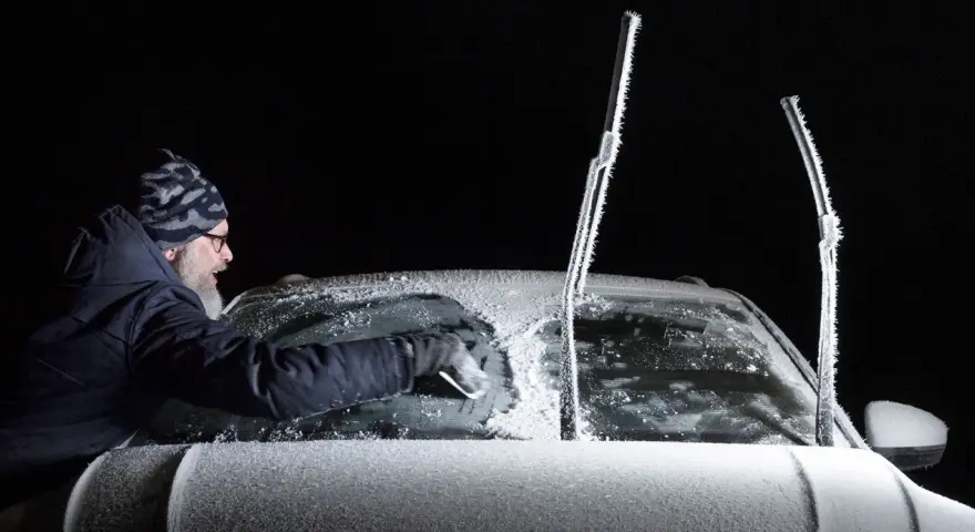 Man skrapar isfri bilruta i mörker. Vindrutetorkarna är täckta av frost.