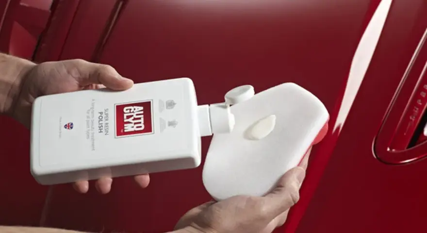 Händer applicerar Autoglym Super Resin Polish på en blank röd bil med en svamp.