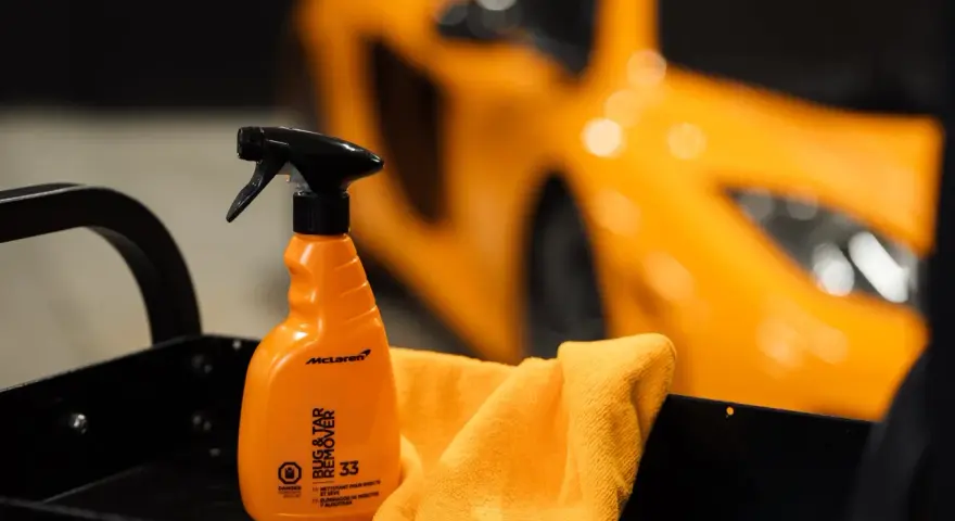 En orange sprayflaska för att ta bort limrester och insekter, bredvid en orange mikrofiberduk. En sportbil i bakgrunden.