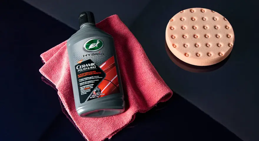 En flaska Turtle Wax Hybrid Solutions Ceramic Polish & Wax, en rosa mikrofiberduk och en polertrissa ligger på en mörkblå yta.