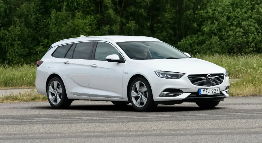 Vit Opel Insignia kombi parkerad vid en grönskande skog. En förare syns i bilen, kanske funderar på ett opel insignia problem.
