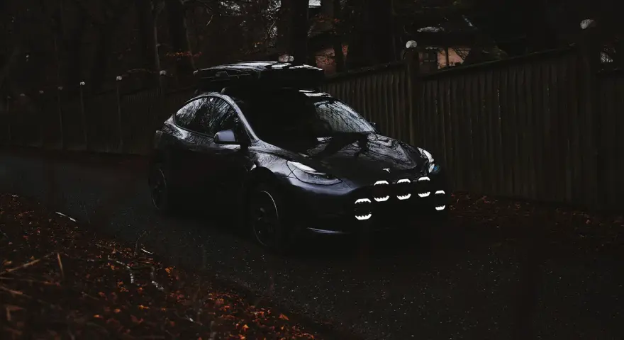 Mörk Tesla med takbox och extraljusstag redo för äventyr längs en höstfärgad väg.