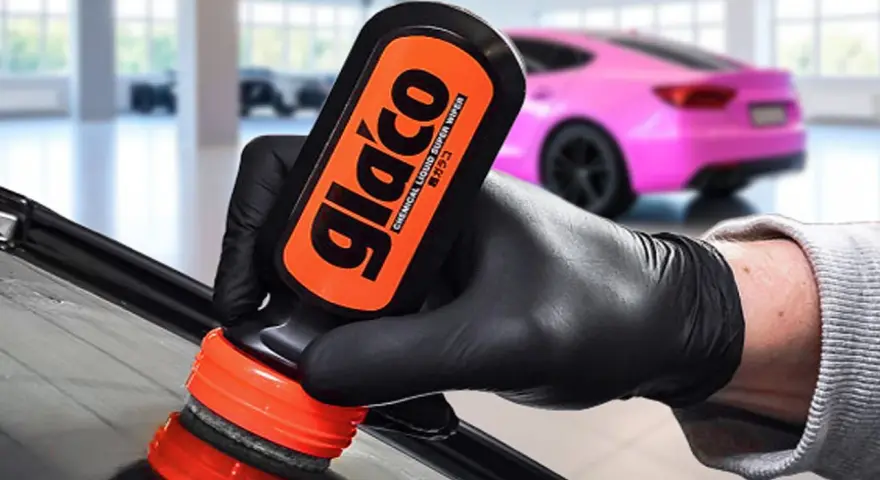 En hand med svart handske applicerar glasförsegling från en Glaco-flaska på en bilruta. En rosa bil syns i bakgrunden.