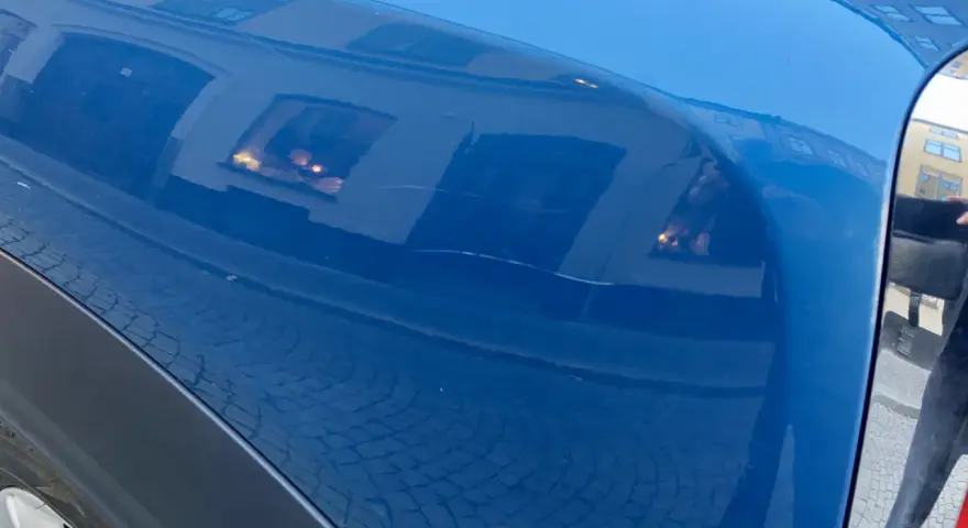 Blå bilskärm med reflektioner av byggnader och kullersten. En repa syns, kanske från kalk borttagning.