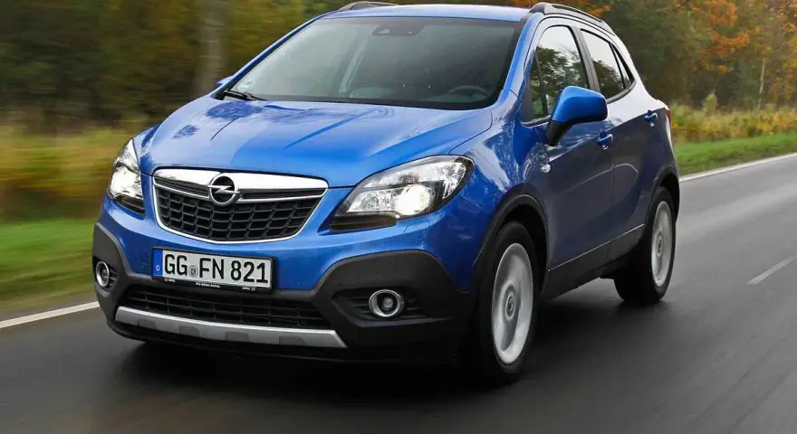 Blå Opel Mokka kör på en väg, kanske med ett opel mokka problem. Träd i höstfärger syns i bakgrunden.