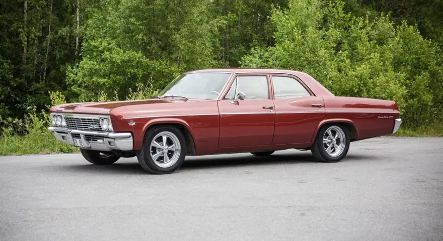 Röd Chevrolet Impala från 1965 med en tung cut, parkerad vid en skogsdunge.