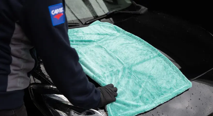 Person i handskar lägger en turkos mikrofiberduk på en svart bilhuv. Duken är från the rag company.