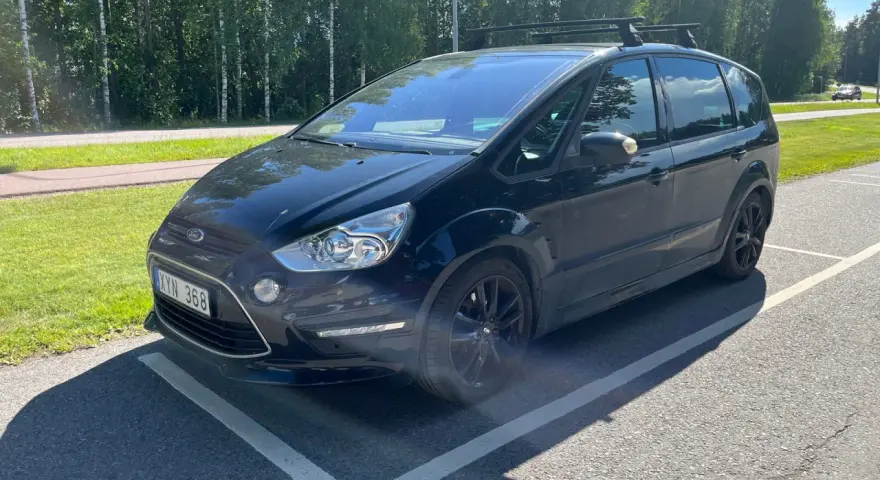 Svart Ford S-Max med takräcke parkerad på en asfalterad yta. En liten bit av stötfångaren verkar vara skadad, ett potentiellt **Ford S-Max problem**.