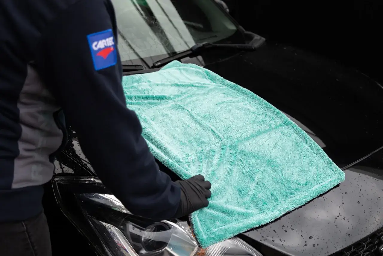En person med svarta handskar lägger en turkos mikrofiberduk på en svart bil. Enkel metod för att vaxa bilen utan att polera.
