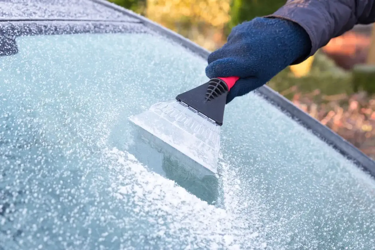 En hand med blå vante skrapar is från en bilruta. Frosten gnistrar i solljuset.