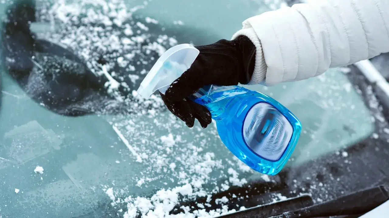 En hand med handske sprayar en bl&aring; v&auml;tska p&aring; en sn&ouml;t&auml;ckt vindruta f&ouml;r att f&aring; isfria bilrutor.