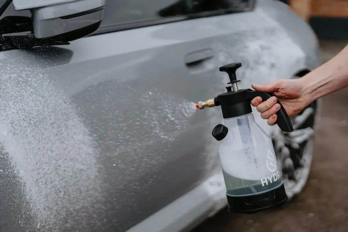 En hand håller en tryckspruta med skum som sprutas mot en grå bil. Sprayflaskor är bra för bilvård.