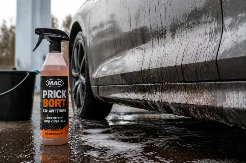 En flaska med MAC Prick Bort kallavfettning står bredvid en smutsig bil. Denna produkt är perfekt för att få bort asfalt, tjära och olja, vilket ger en renare bil.