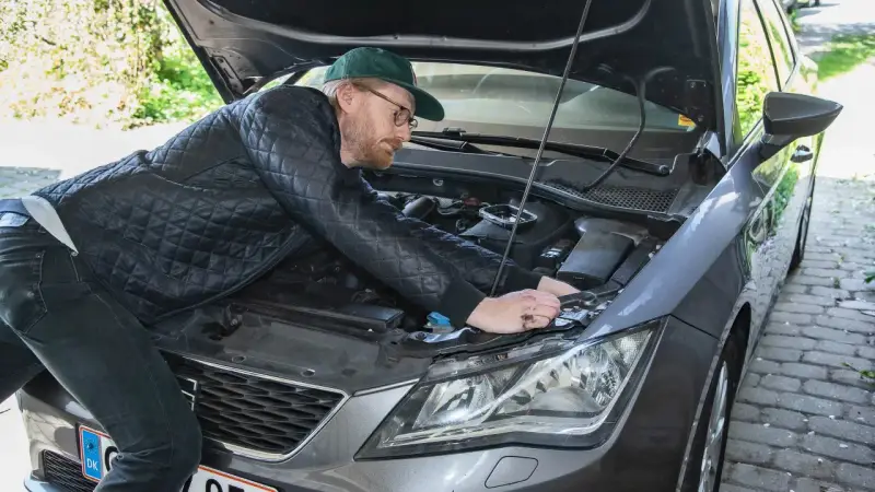 Man med keps och glasögon mekar med bilens motor, kanske byter en h7 led-lampa.